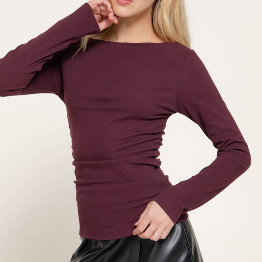 A woman modeling a long-sleeve, boat-neck ribbed top in deep burgundy, paired with a black faux-leather mini skirt, against a plain off-white background.