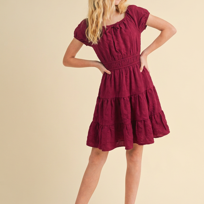 A young woman with blonde hair models a short-sleeved, tiered burgundy dress against a solid beige studio background. She is wearing white woven flat shoes.
