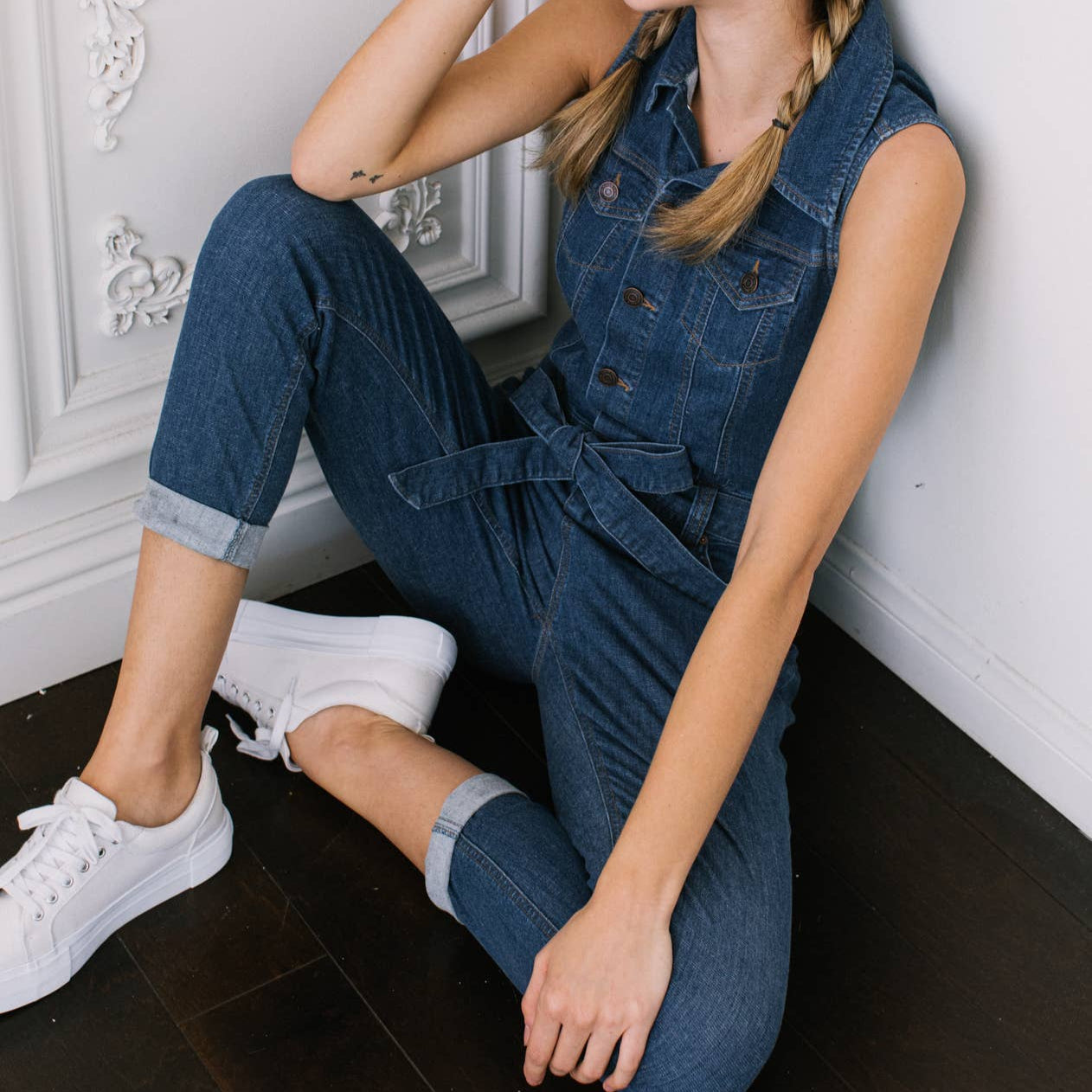 Woman wearing a denim jumpsuit sitting on the floor against a white wall.