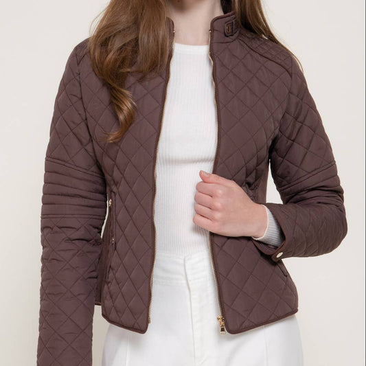 A woman wearing a brown diamond-quilted cropped jacket over a white top and white pants, photographed against a light neutral studio background. A small black 'PLUS SIZE' label appears in the top-right corner.