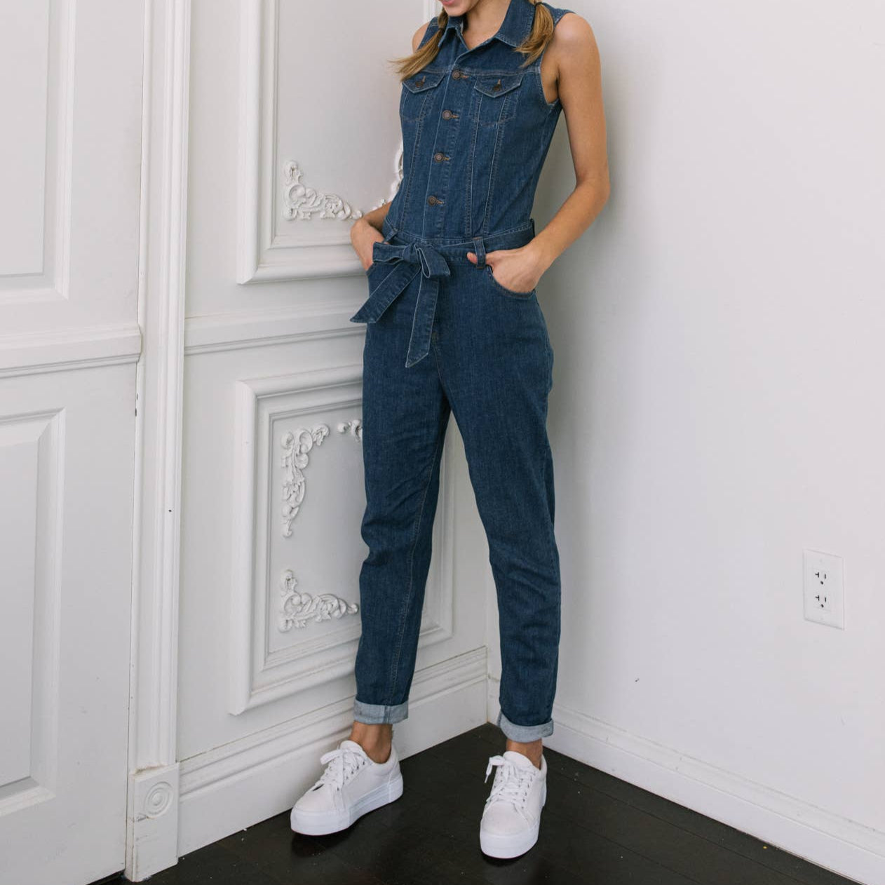 Woman wearing a denim jumpsuit standing against a white wall.
