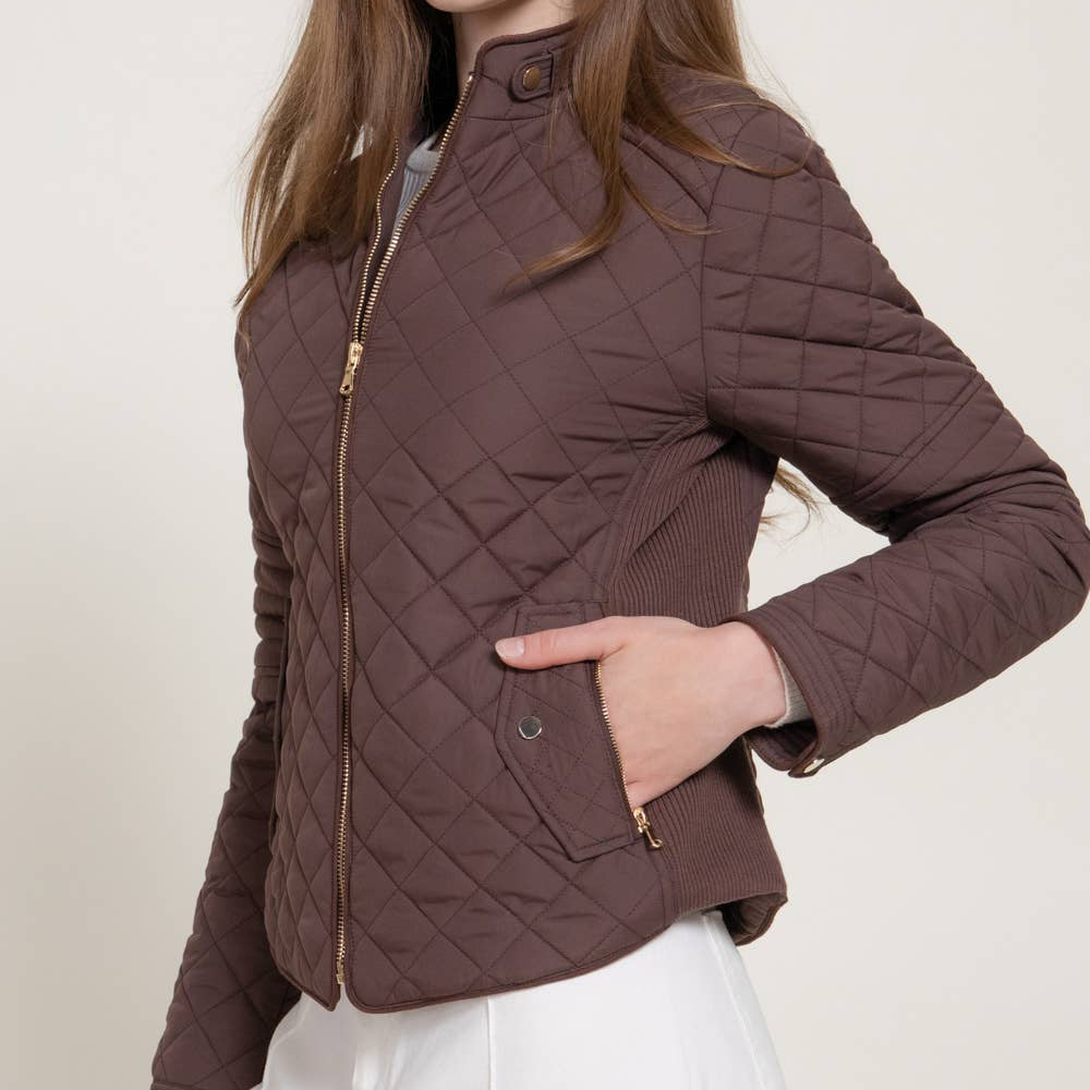 A woman wearing a brown diamond-quilted zip-up jacket with gold-tone hardware, paired with white pants, against a light beige studio background. A black 'PLUS SIZE' label is in the top right corner.