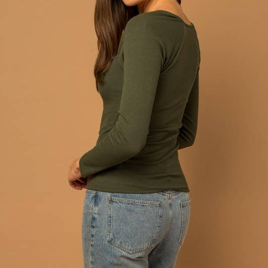 Woman wearing an olive green long-sleeve top and blue jeans against a brown background