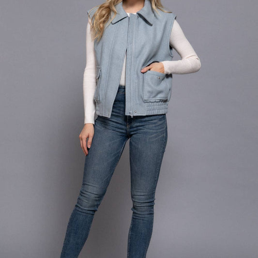 Woman wearing a light blue denim vest over a white shirt, paired with blue jeans and black boots on a gray background.