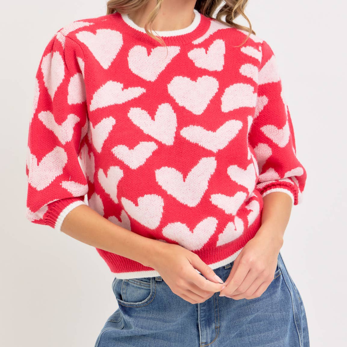 Woman wearing a red sweater with white heart patterns and blue jeans on a light gray background