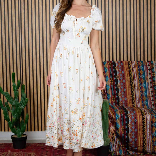 Woman in a white floral dress standing against a striped wall with a colorful rug and plant.