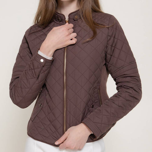 A woman wearing a brown diamond-quilted zip-up jacket with white pants, posed against a light beige studio background. A black 'PLUS SIZE' label is visible in the top right corner.