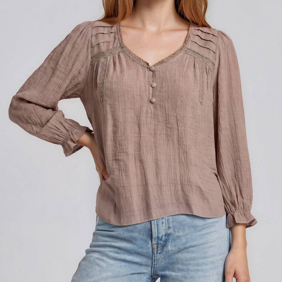 A woman modeling a long-sleeve, light brown linen blouse with pleated details on the chest and a buttoned henley neckline, paired with light blue jeans, against a solid light gray background.
