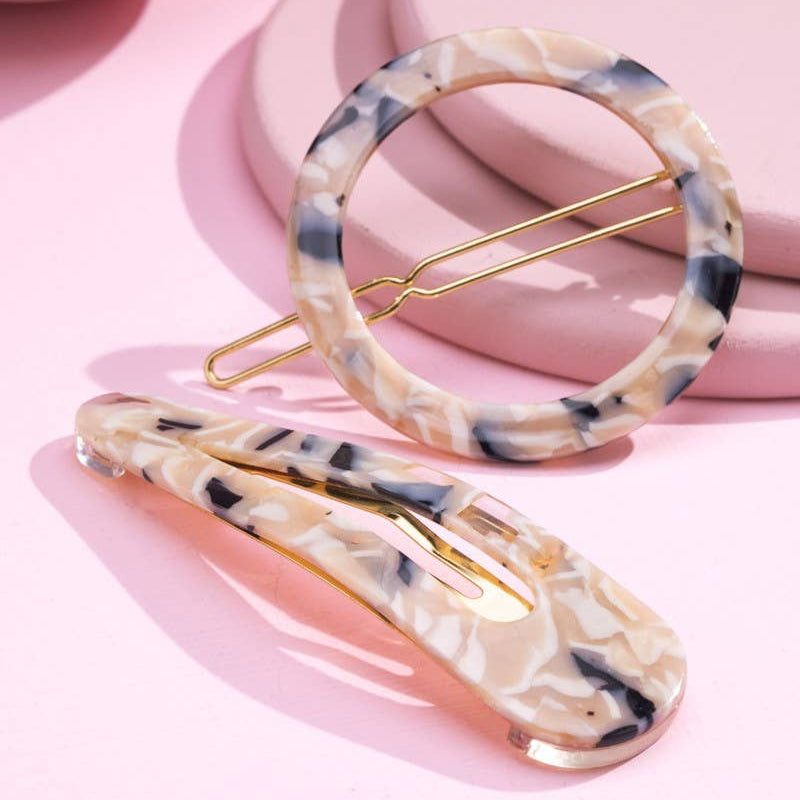Two tortoiseshell hair clips with gold-tone metal clasps, displayed on a pink background with soft pink props.