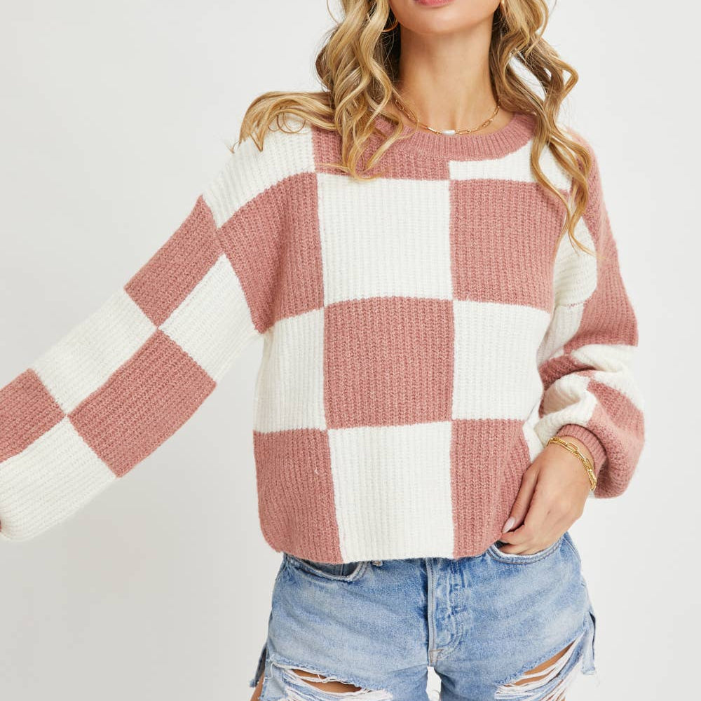 A woman wearing a pink and white checkered knit sweater with long sleeves, paired with distressed denim shorts, against a light gray studio background.