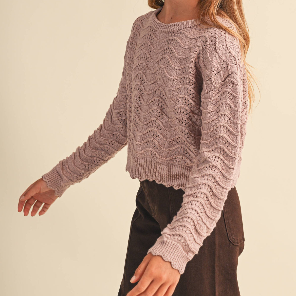 A young woman modeling a dusty mauve knit sweater with a textured wavy pattern and scalloped edges, paired with dark brown pants, against a solid light beige background.