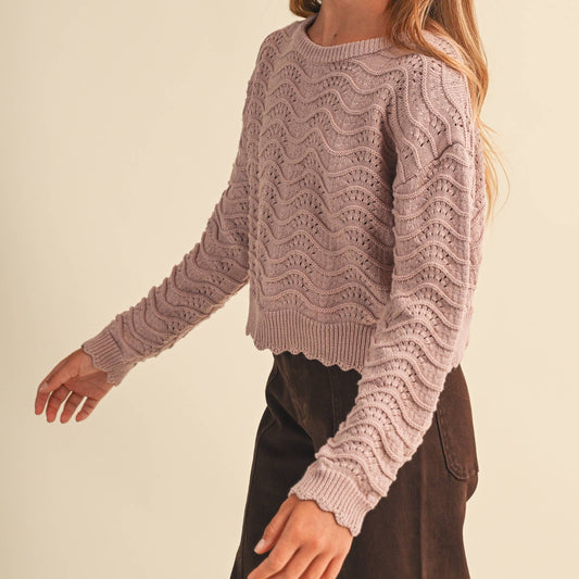 A young woman modeling a dusty mauve knit sweater with a textured wavy pattern and scalloped edges, paired with dark brown pants, against a solid light beige background.