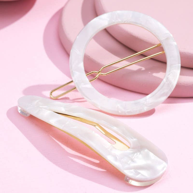 Two white mother-of-pearl hair clips with gold-tone metal spring clips, arranged on a pink background with soft pink geometric props.
