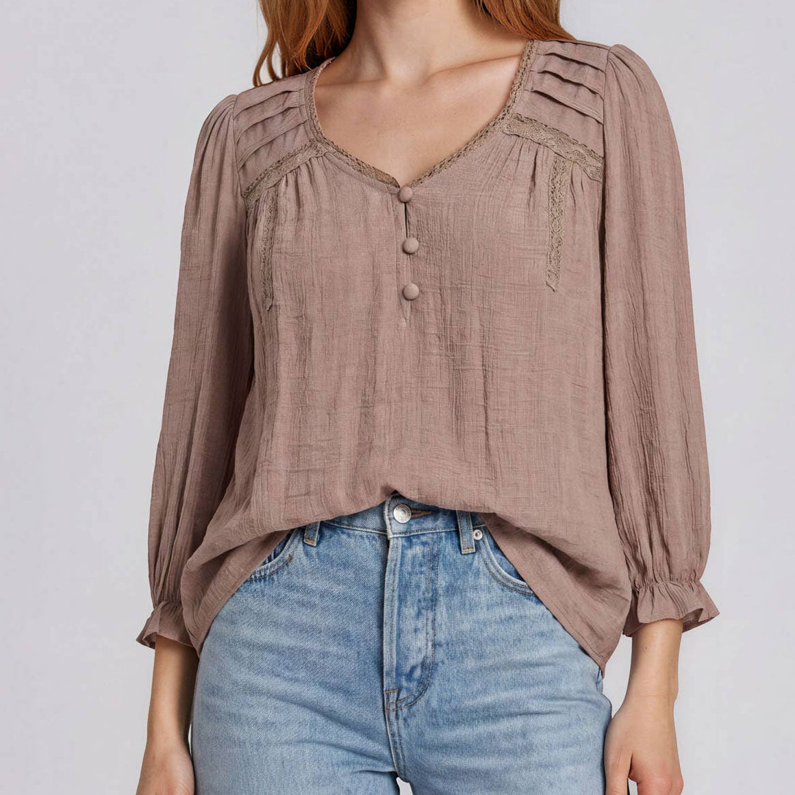 A woman modeling a taupe-brown long-sleeve blouse with decorative shoulder straps and a buttoned V-neck, paired with light-wash blue jeans against a light gray studio background.