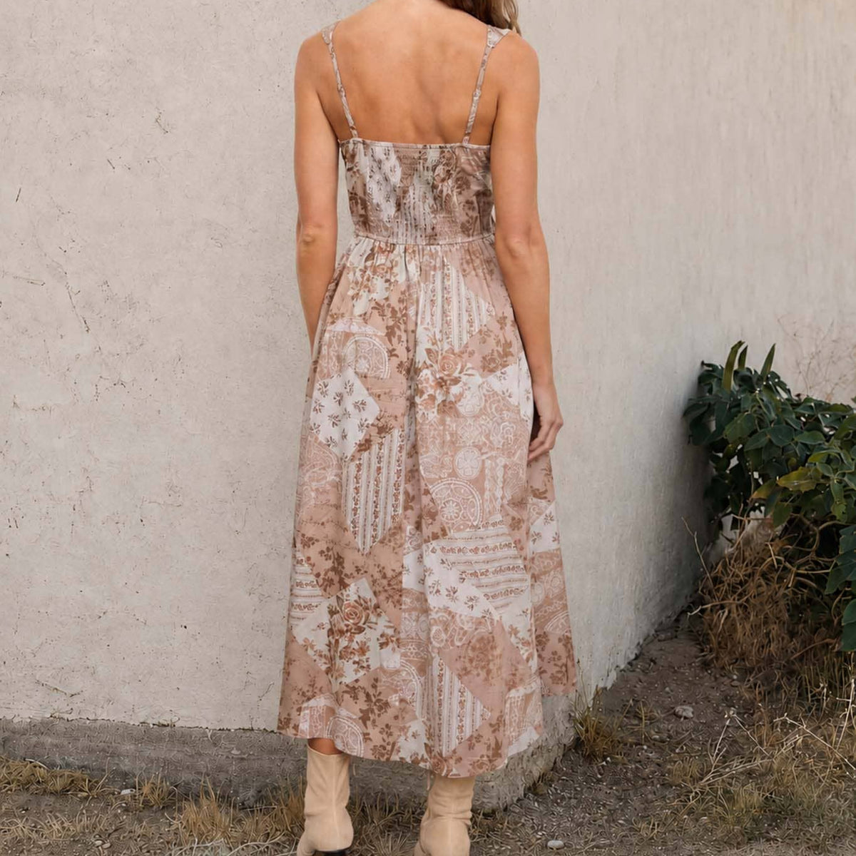A woman viewed from behind wearing a sleeveless midi dress with a beige and brown patchwork floral pattern, standing outdoors against a textured stucco wall.
