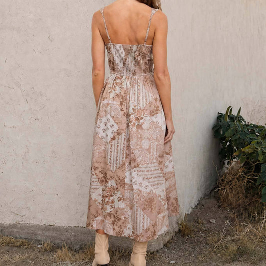 A woman viewed from behind wearing a sleeveless midi dress with a beige and brown patchwork floral pattern, standing outdoors against a textured stucco wall.