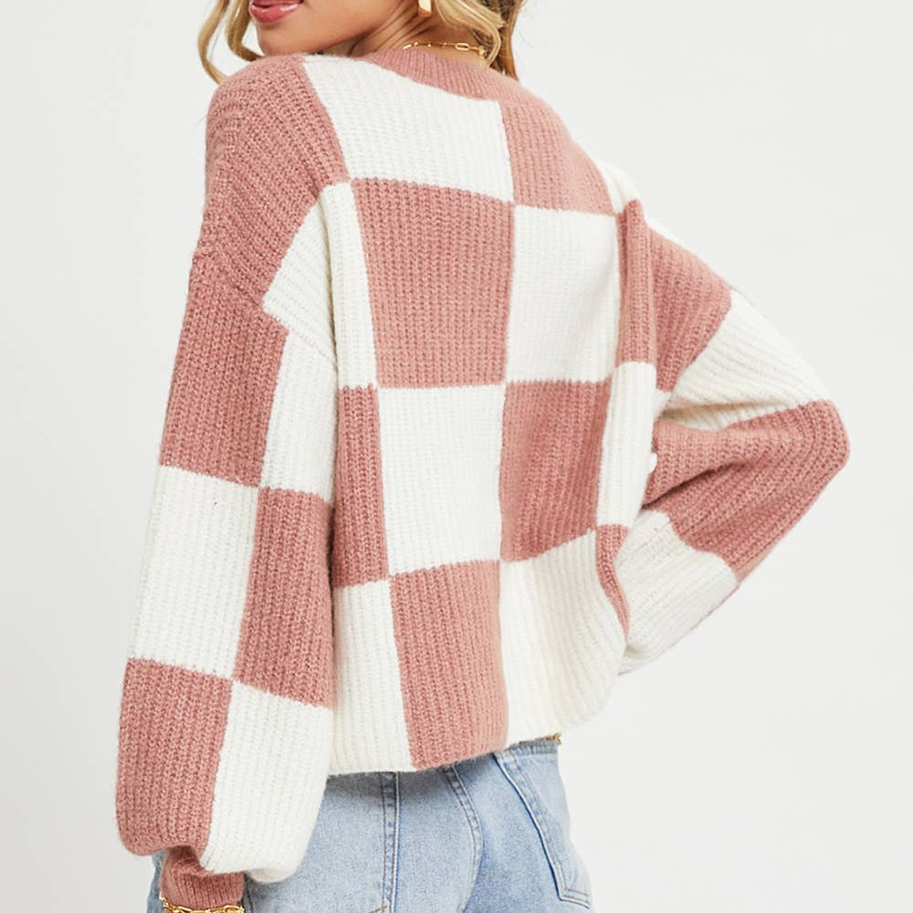 A woman wearing a pink and white checkered knit sweater with light blue denim shorts, photographed against a plain light gray studio background.