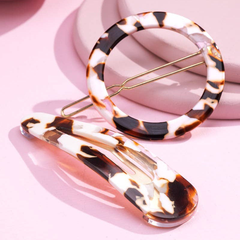 Two tortoiseshell hair clips on a pink background, one circular ring clip and one elongated oval clip, with gold-tone metal spring clips.