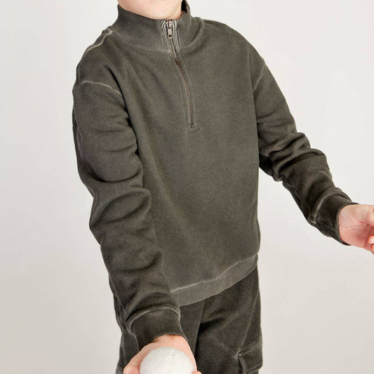 A young boy wearing a dark green quarter-zip fleece pullover and a black-and-white baseball cap, holding a white ball against a light gray studio background.