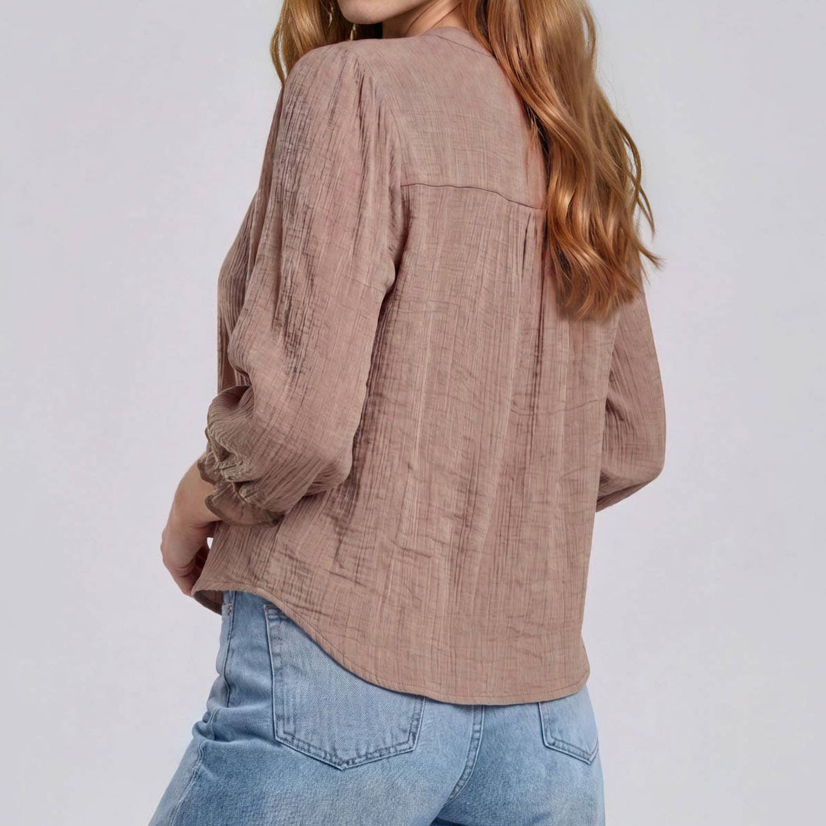 A woman with long light brown hair is shown from a back-three-quarter angle wearing a textured, dusty rose long-sleeve blouse and light blue jeans against a plain light gray studio background.