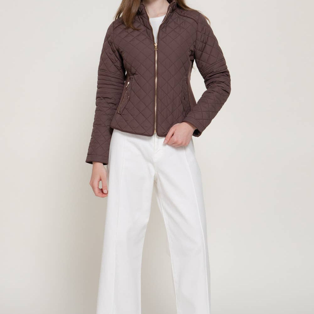 A model wearing a dark brown quilted jacket, white wide-leg trousers, and black boots, standing against a plain light beige studio background.
