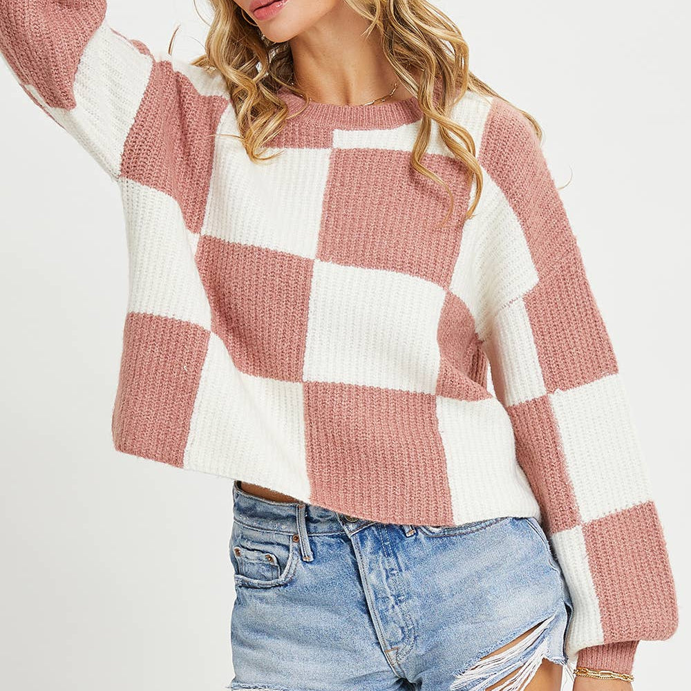 A woman wearing a pink and white checkered knit sweater with distressed denim shorts, posing against a light gray studio background.