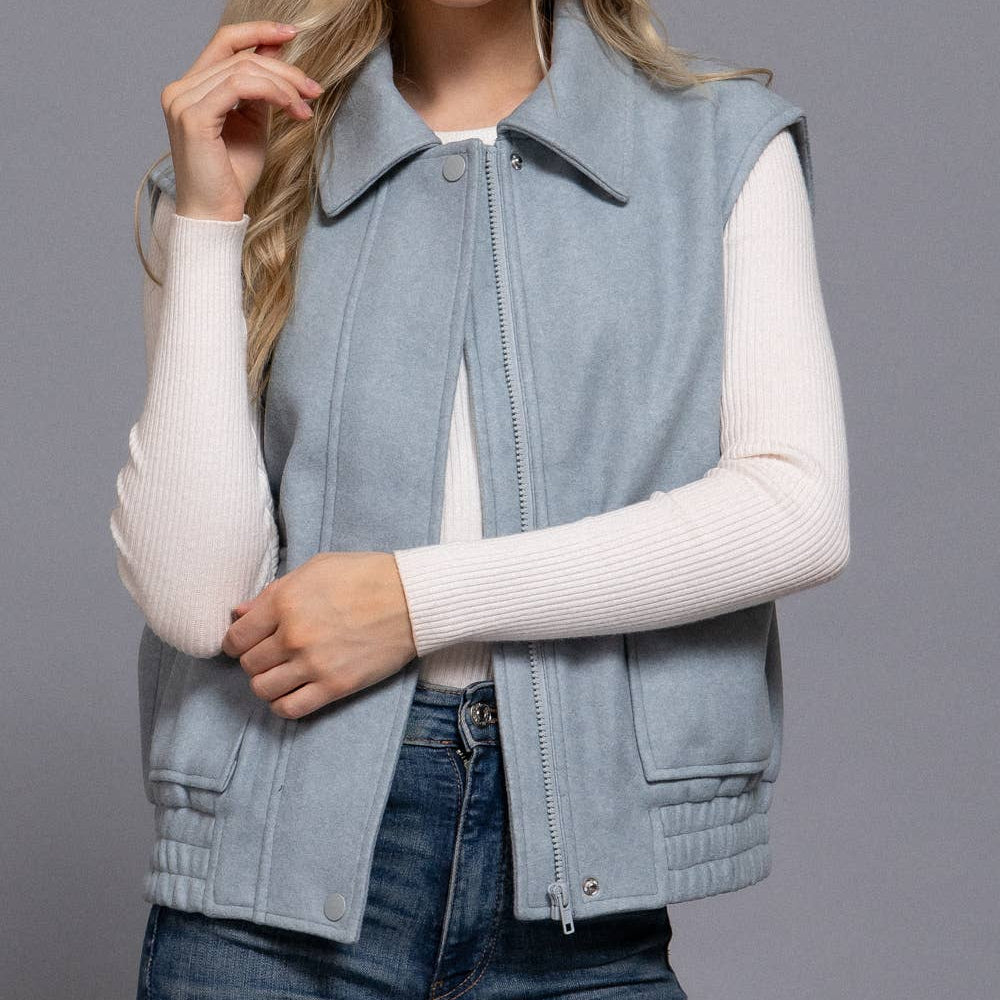 Woman wearing a light blue vest over a white sweater and jeans on a gray background