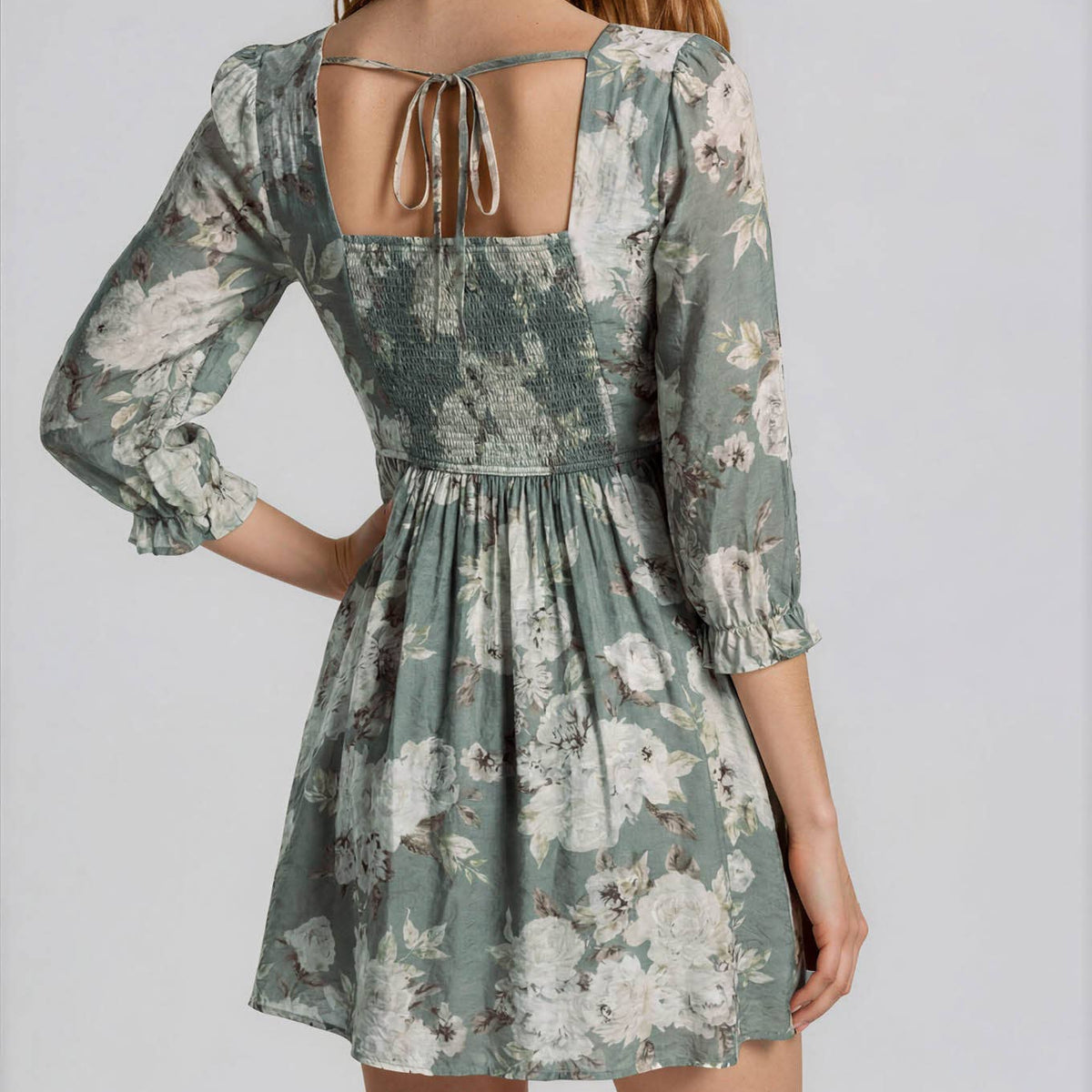 A woman modeling a three-quarter sleeve floral dress from the back, featuring a tie-back neckline and a flared skirt, set against a plain light gray studio background.