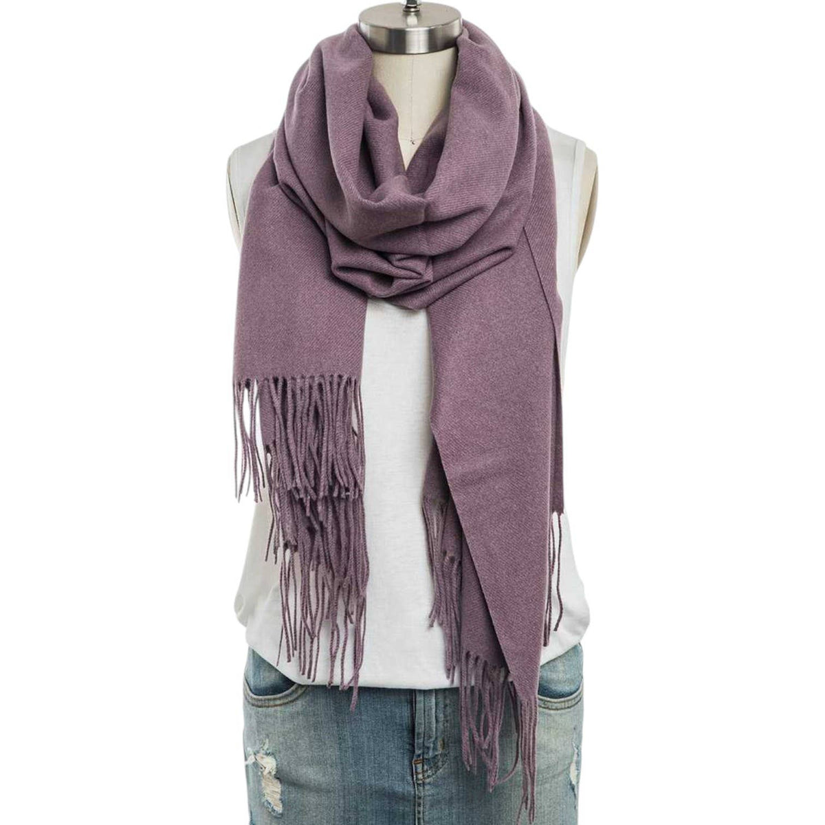 A dusty purple knit scarf with fringe trim, styled on a dress form mannequin wearing a white sleeveless top and blue jeans against a plain white background.