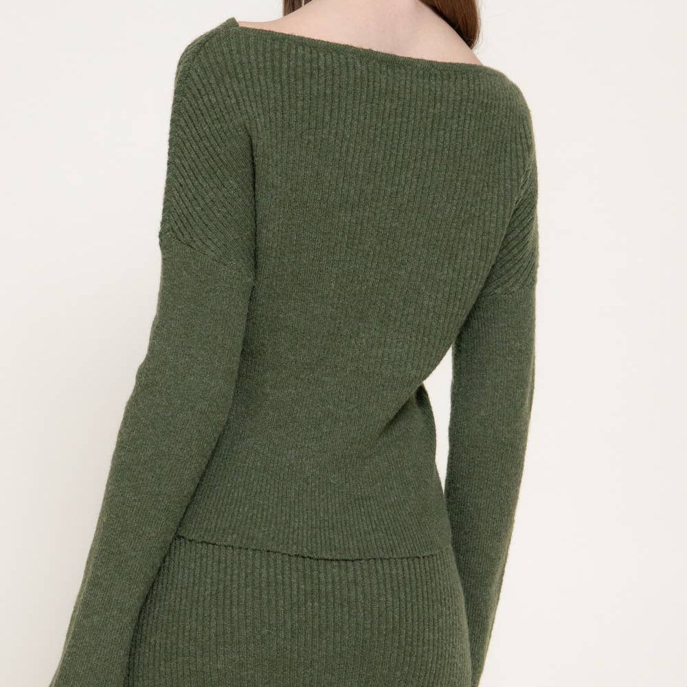 A woman wearing an olive green ribbed knit sweater and matching skirt, shown from a side-back angle against a plain off-white studio background.
