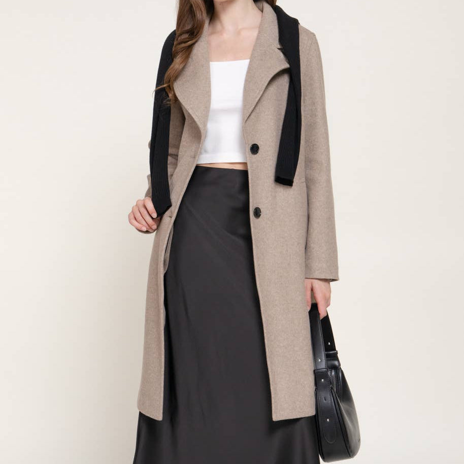 A woman models a long beige coat with black lapels and black buttons, worn over a white top and a long black skirt, holding a black handbag against a plain light beige studio background.