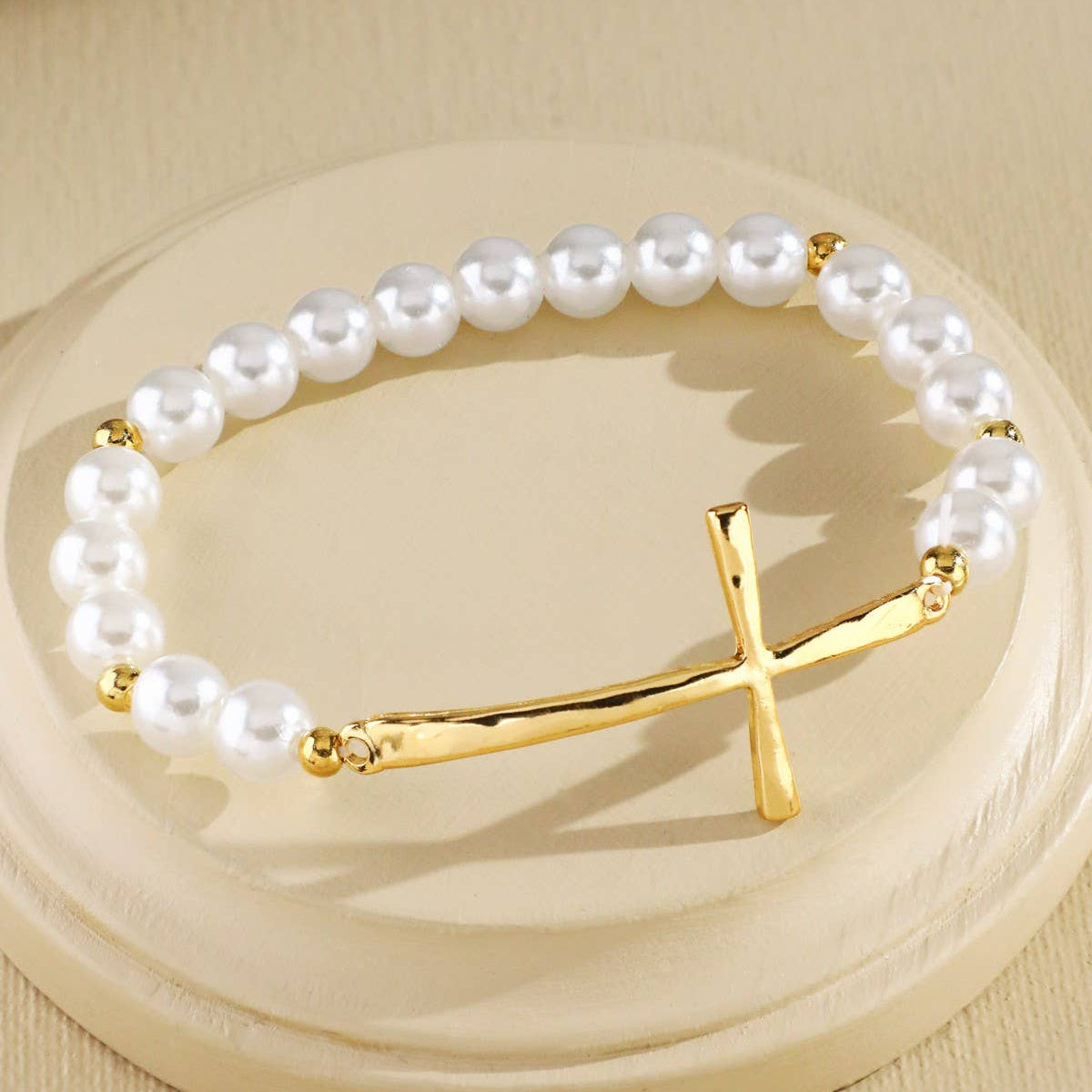 A pearl-bead bracelet with a polished gold cross clasp, displayed on a round beige pedestal against a matching beige background.