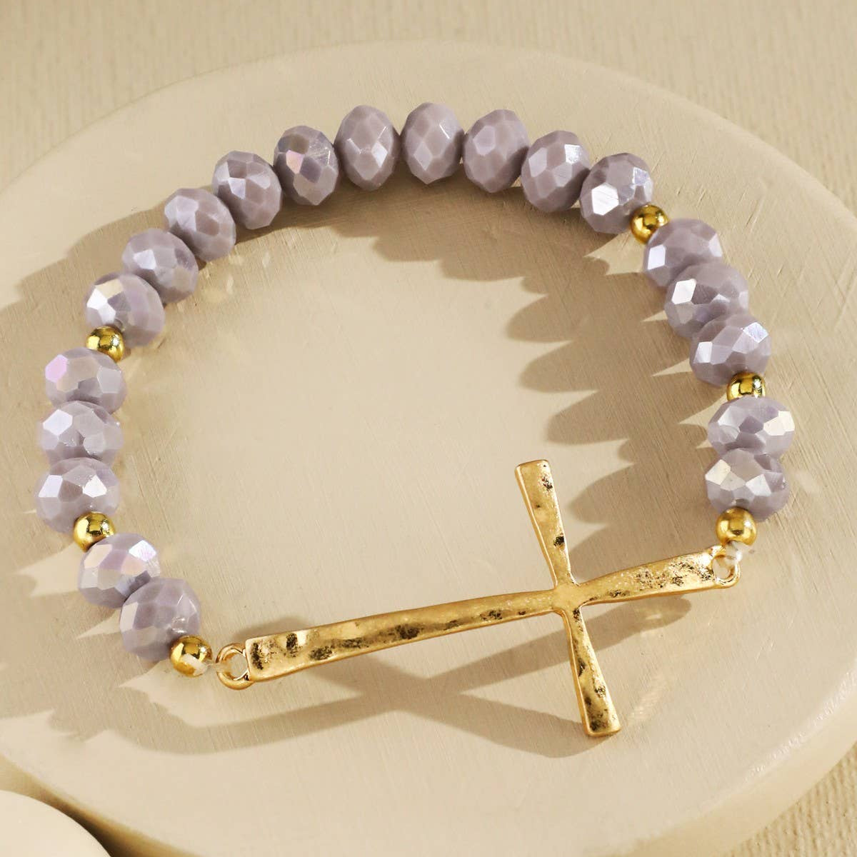 A beaded bracelet with faceted lavender-gray beads and a textured gold cross charm, displayed on a round beige pedestal.