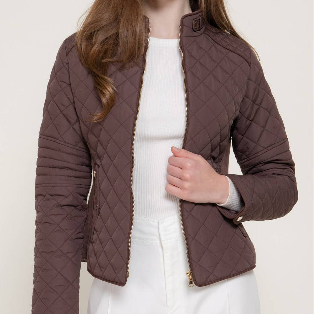 A woman wearing a brown diamond-quilted cropped jacket over a white top and white pants, photographed against a light neutral studio background. A small black 'PLUS SIZE' label appears in the top-right corner.