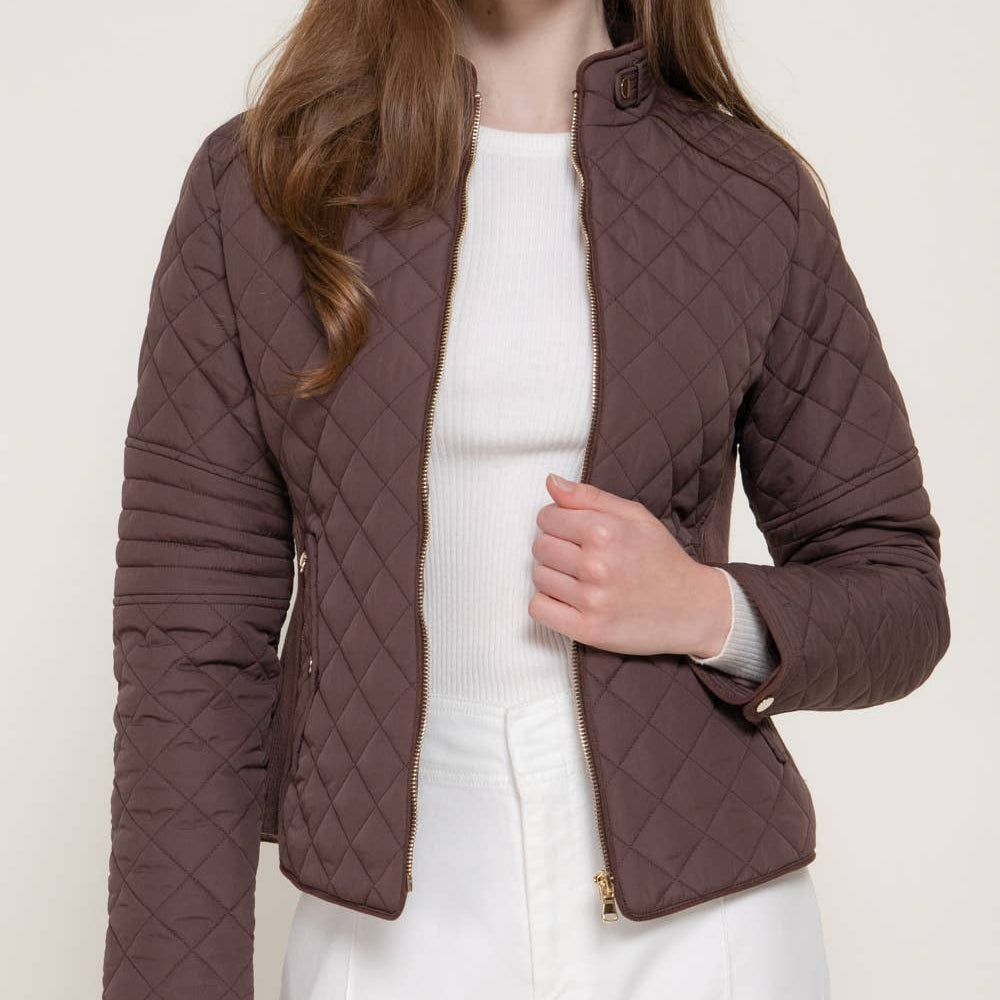 A woman modeling a short, quilted jacket in a dark brown color, worn over a white ribbed top and white pants, against a light beige studio background.