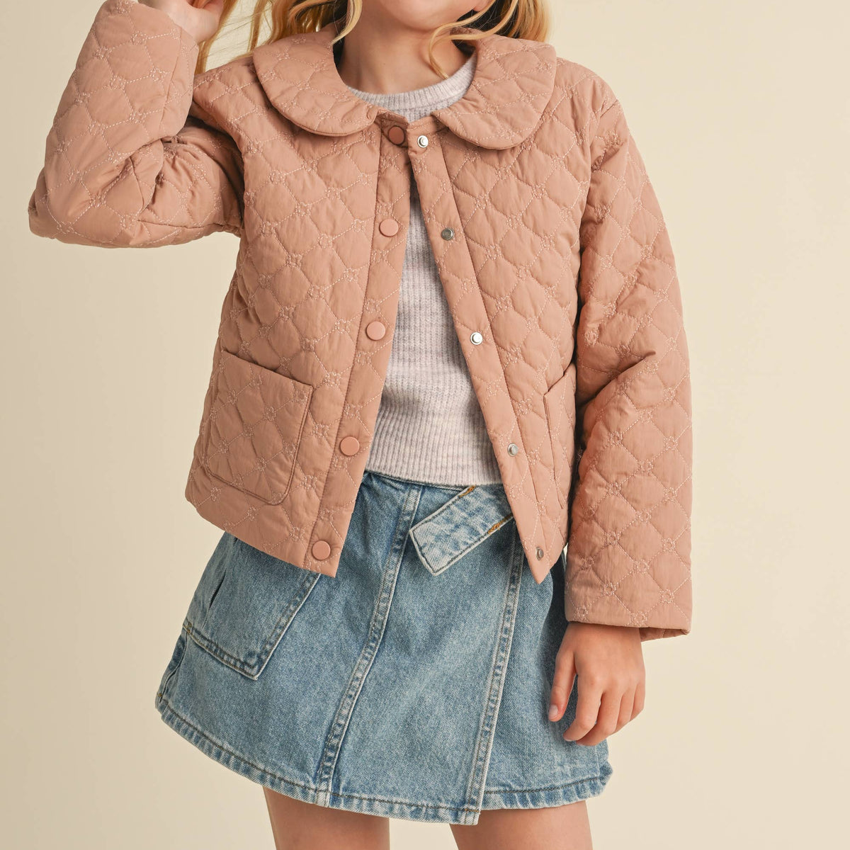 A young girl with blonde hair wearing a dusty pink quilted jacket, a light gray ribbed sweater, and a denim mini skirt, posing against a solid beige background.