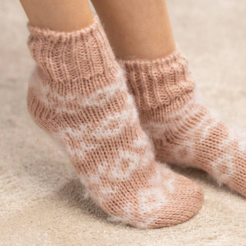 A close-up of a person's feet wearing fuzzy knit ankle socks in a dusty pink and white pattern, standing on a soft beige carpet.