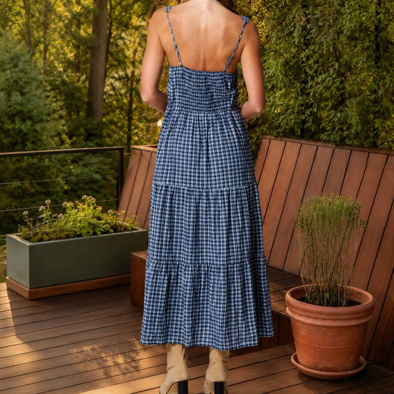 Woman in a blue checkered dress standing on a wooden deck with plants and greenery.