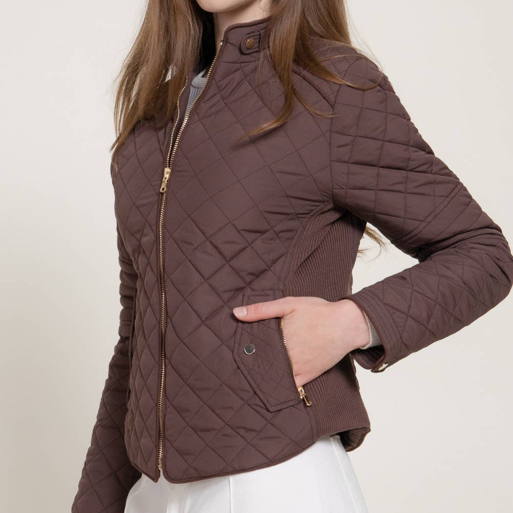 A woman wearing a brown diamond-quilted jacket with gold-tone zippers, paired with white pants, photographed against a plain light beige studio background.