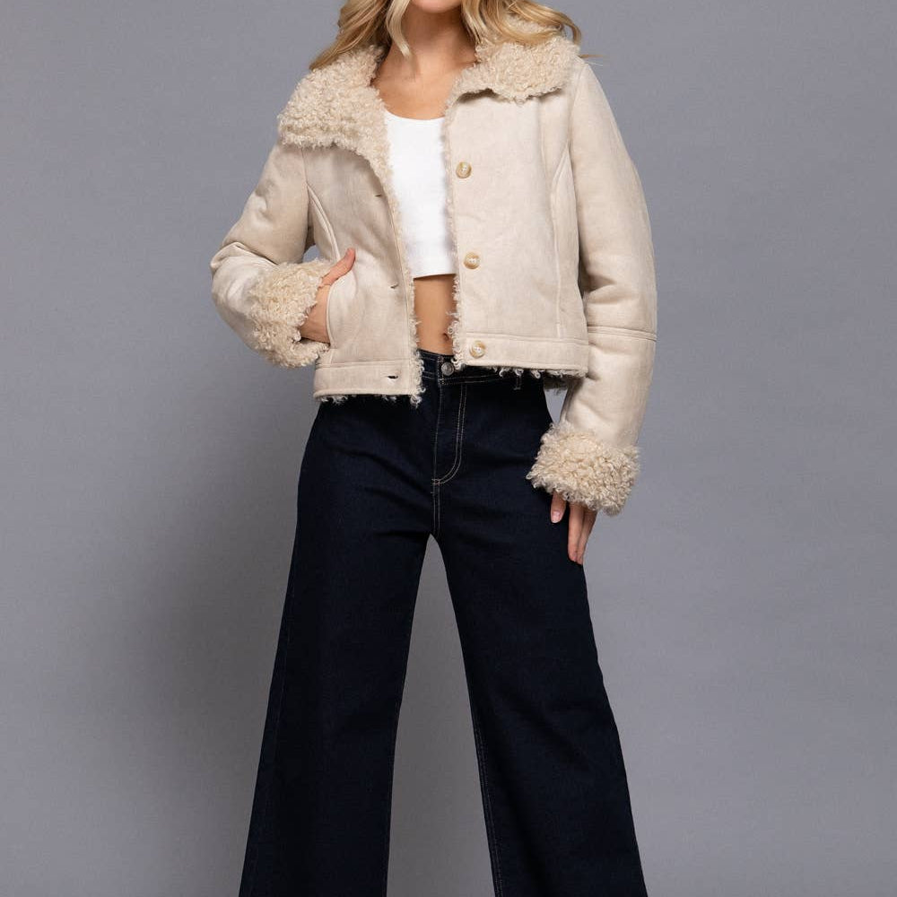 Woman wearing a beige jacket with fur trim, white top, and black pants on a gray background