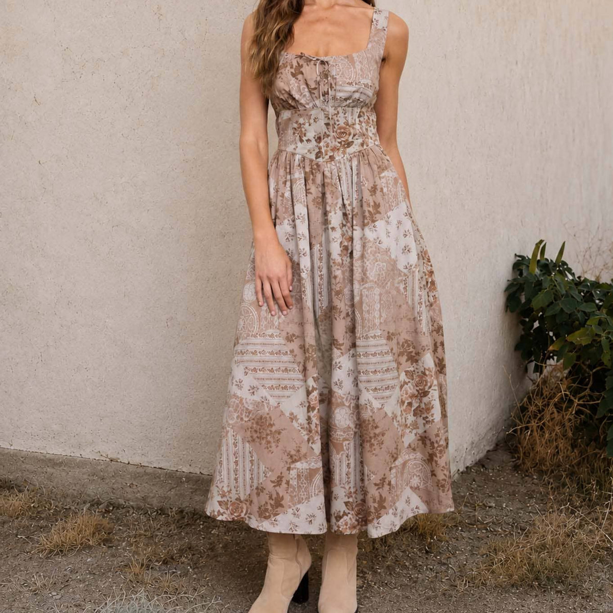 A woman stands outdoors against a stucco wall wearing a sleeveless midi dress with a beige and brown floral and geometric pattern, paired with beige ankle boots.