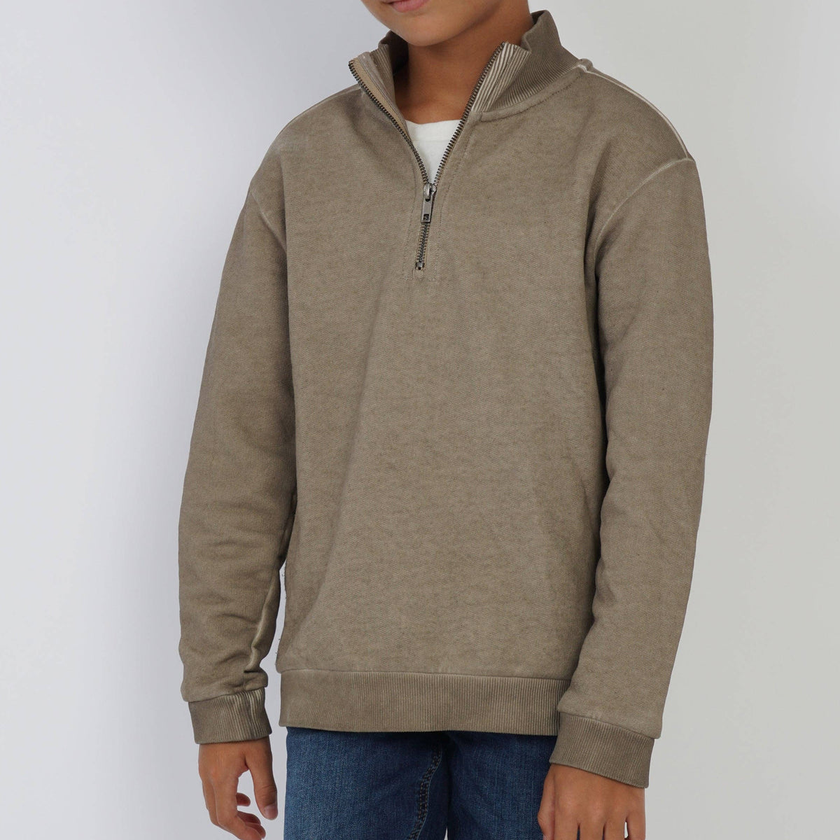 A young boy wearing a heather taupe-brown quarter-zip pullover sweatshirt with a mock neck, styled over a white t-shirt and blue jeans, standing against a light gray studio background.