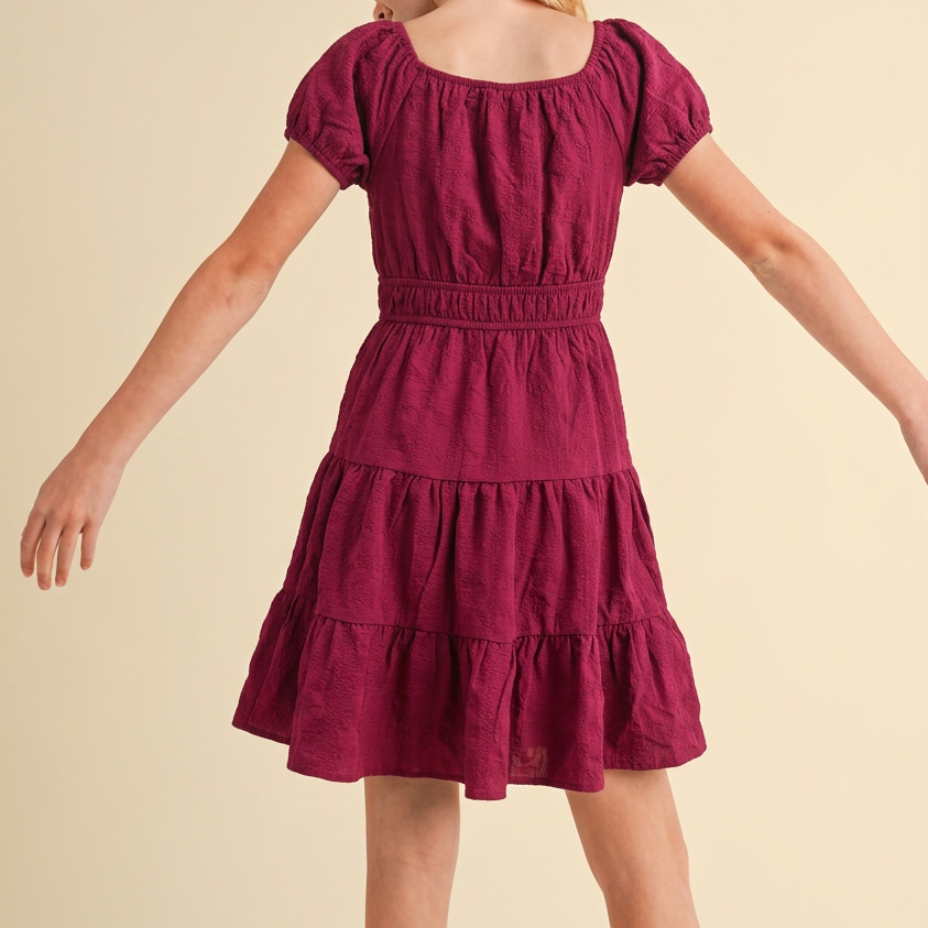 A young girl with blonde hair, viewed from behind, wearing a short-sleeved tiered dress in a deep berry purple color, set against a solid light beige background.