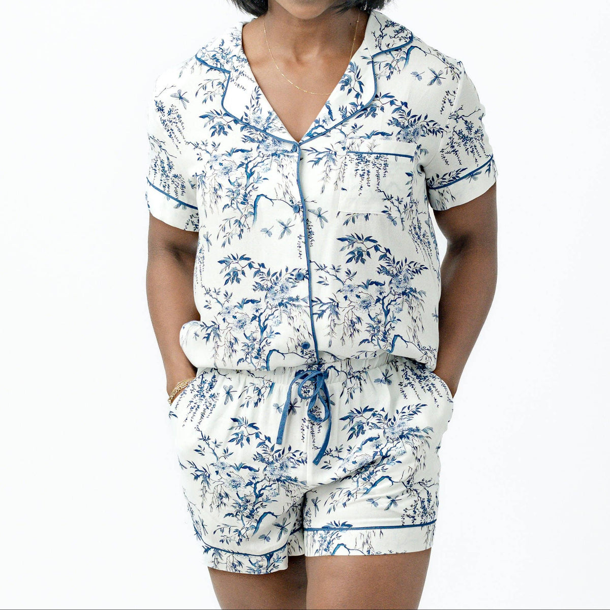 A woman modeling a matching short-sleeve button-front pajama top and shorts set with a blue toile-style landscape print on a white background, photographed against a plain white studio backdrop.