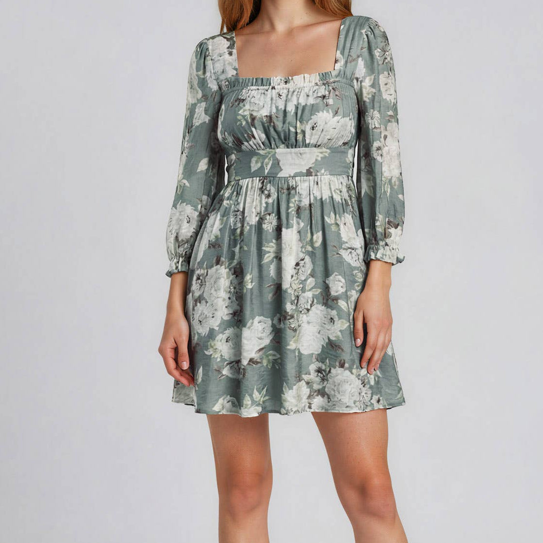 A woman modeling a long-sleeve mini dress with a square neckline and a green-gray floral print, paired with beige high heels, against a light gray studio background.