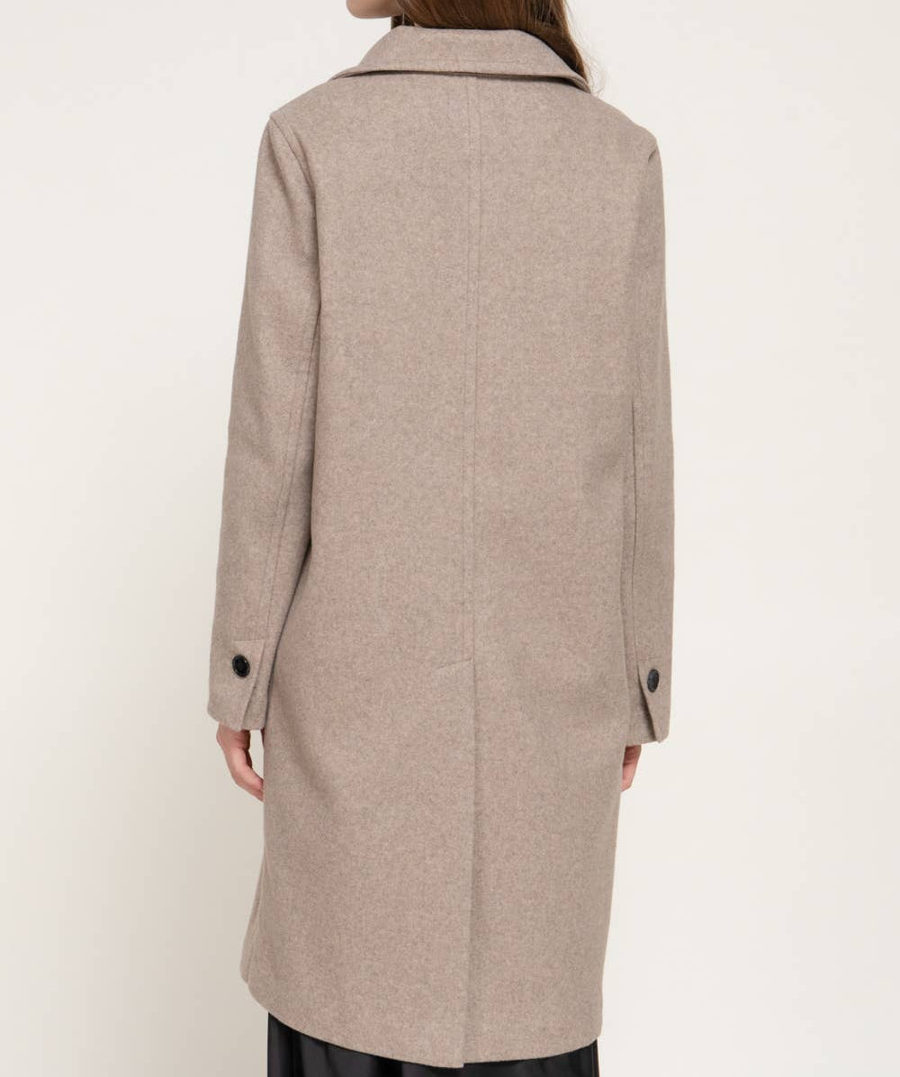 A woman shown from the back wearing a mid-length taupe-beige wool coat with black buttons on the cuffs, paired with black trousers, against a plain off-white background.