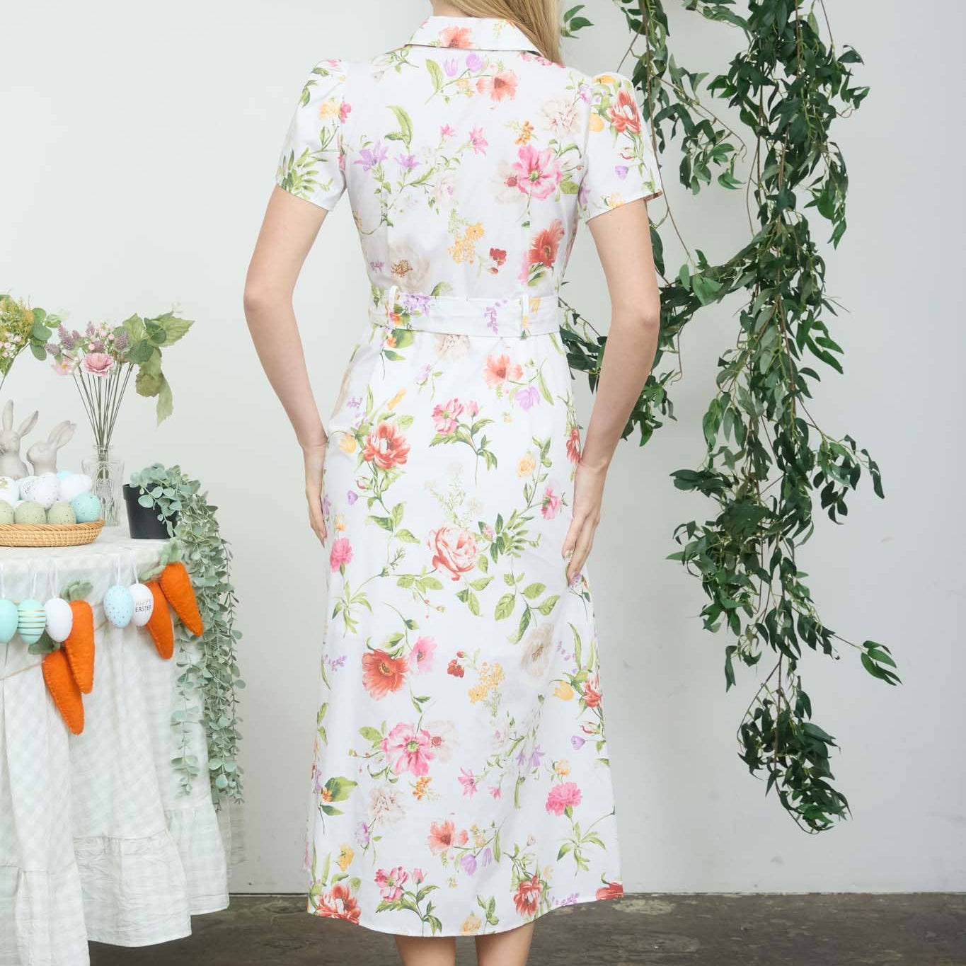Woman wearing a floral dress standing in a room with greenery and a table.