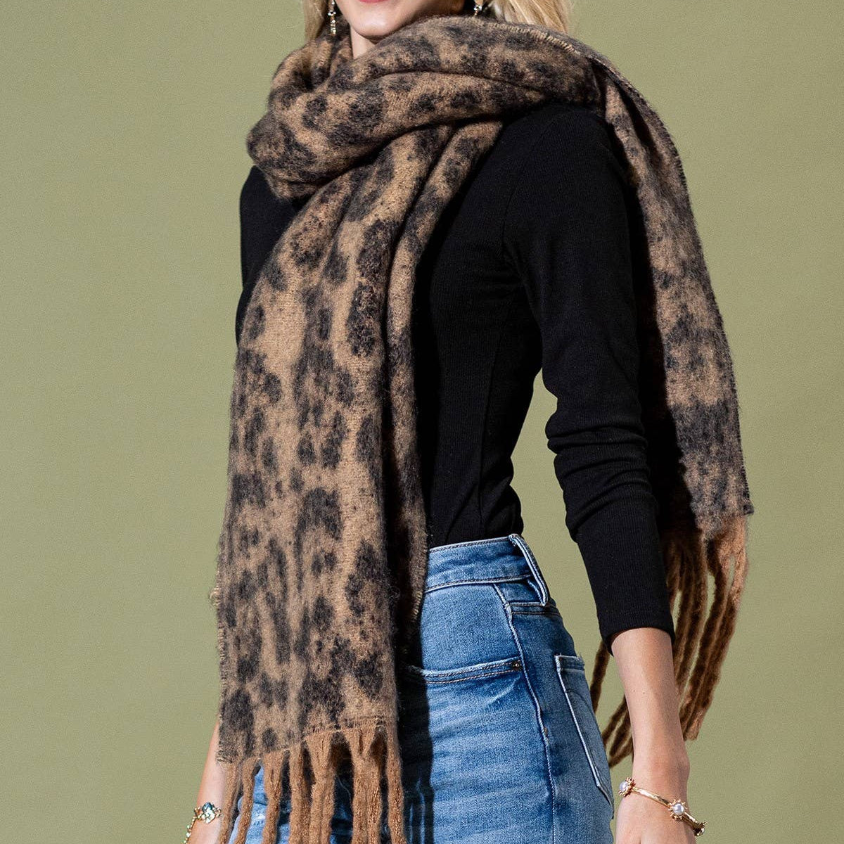 A woman modeling a fuzzy leopard-print scarf with brown fringe, styled over a black long-sleeve top and blue jeans against a solid olive green background.