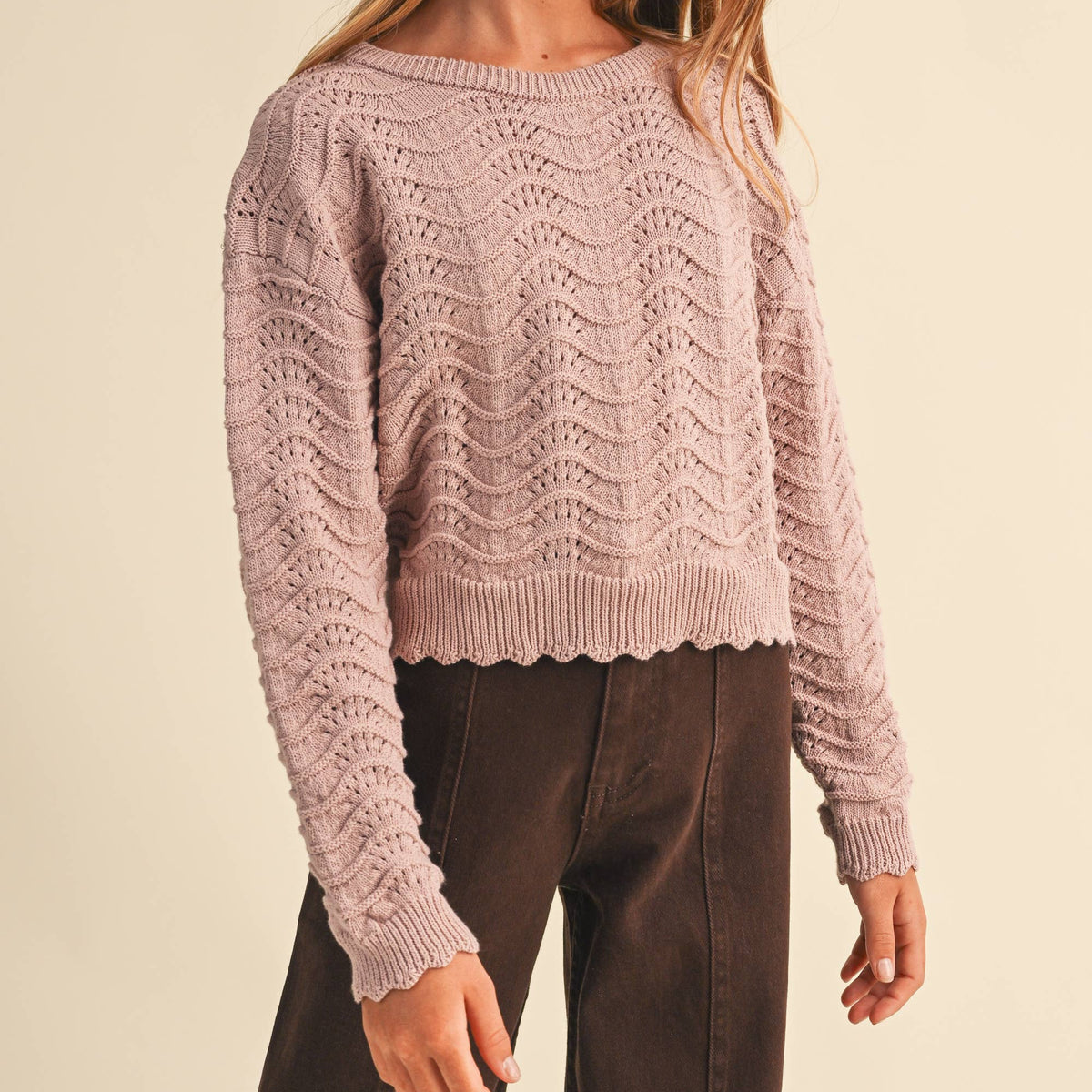 A young girl with long light brown hair wearing a dusty pink cable-knit cropped sweater and dark brown pants, standing against a solid beige studio background.