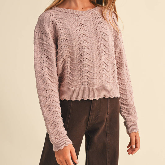 A young girl with long light brown hair wearing a dusty pink cable-knit cropped sweater and dark brown pants, standing against a solid beige studio background.